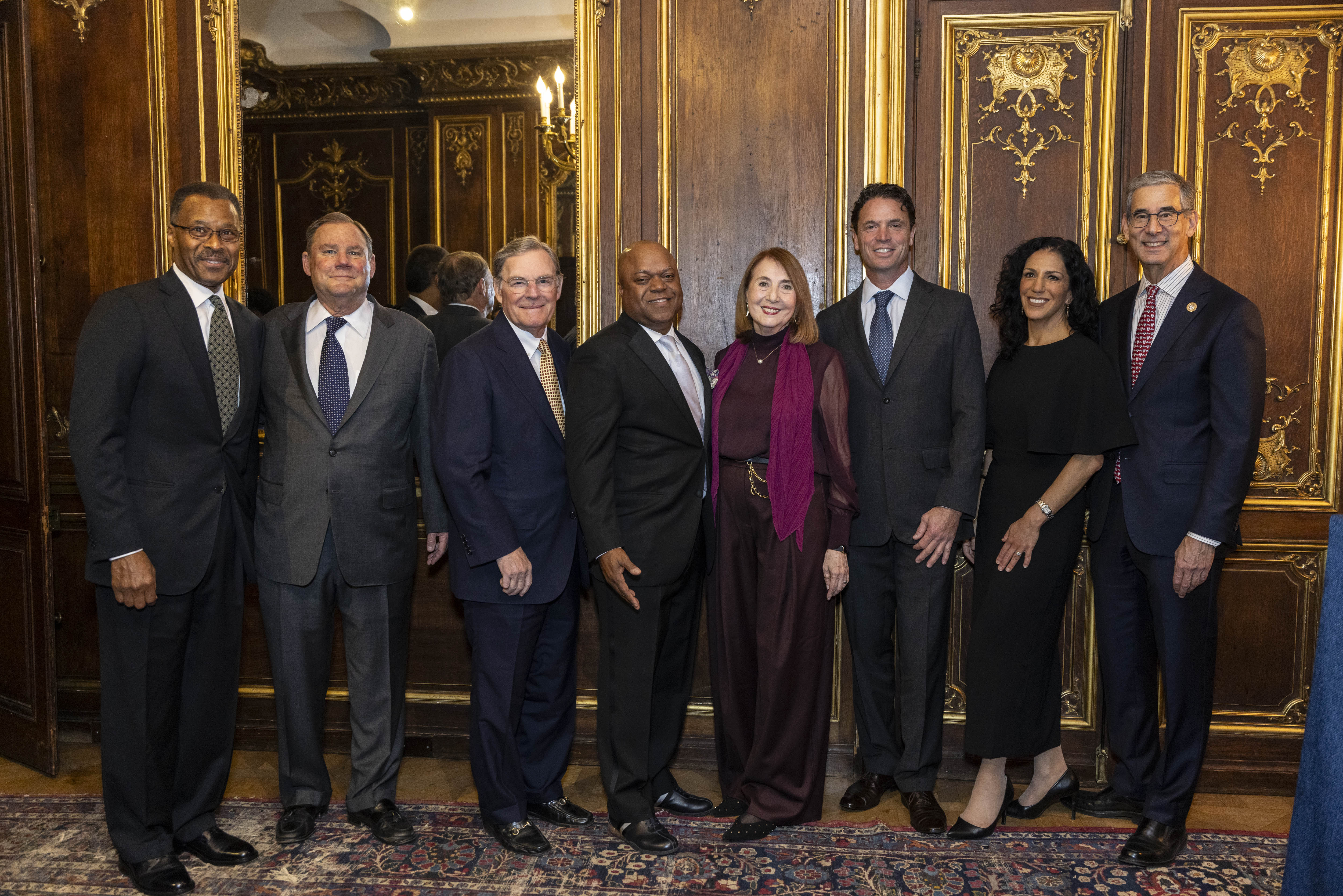 Eight formally dressed adults stand side by side posing for a photo in an ornate room with dark wood-paneled walls, gold decorative detailing, and mirrored panels at the McGraw Prize Gala in 2025.