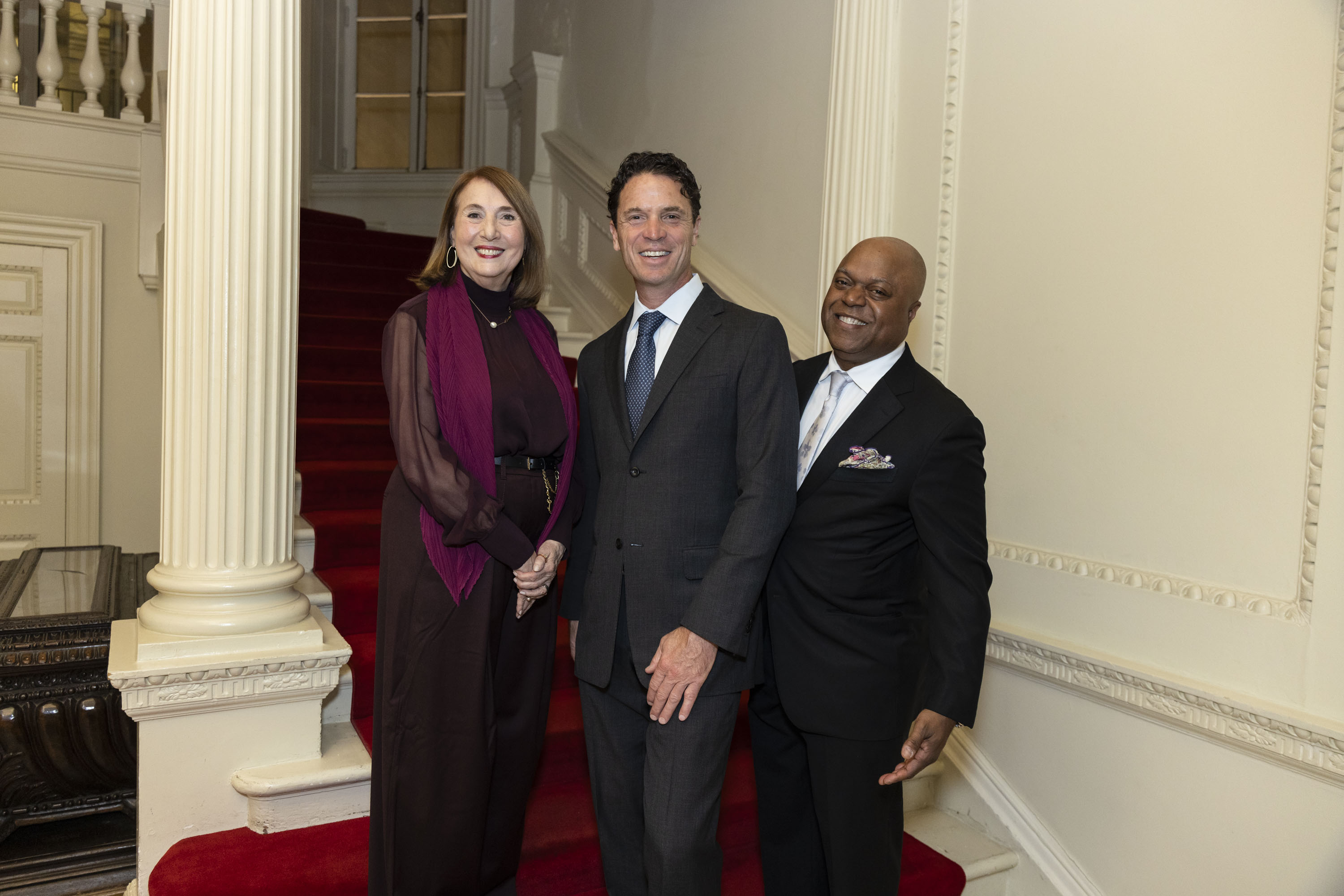 Cathy Davidson, Joe Wolf, and Frederic Bertley standing while smiling against red stairs