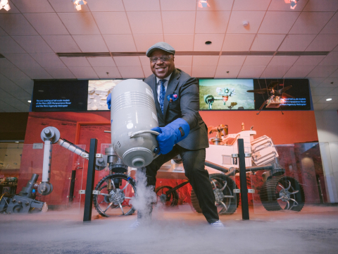 Frederic Bertley wears goggles and mits as he pours out a gray jar with smoke coming out of it at a science exhibit.